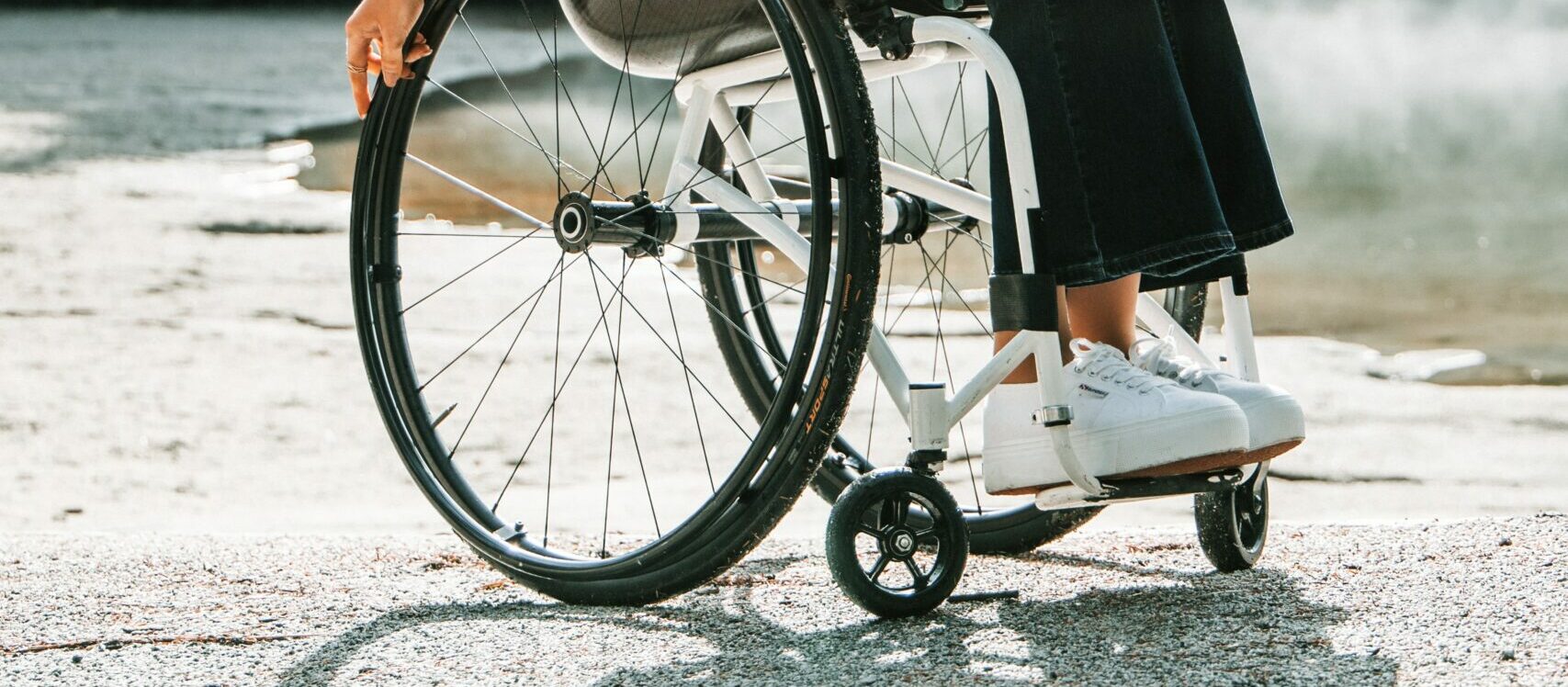 der untere Teil eines Rollstuhls auf einem steinigen Untergrund, der Rollstuhl ist weiß, Greifreifen und Reifen schwarz. Eine Frau sitzt im Rollstuhl, zu sehen sind die Beine. Sie trägt eine Jeans und weiße Schuhe. Die Füße stehen auf dem Fußbrett